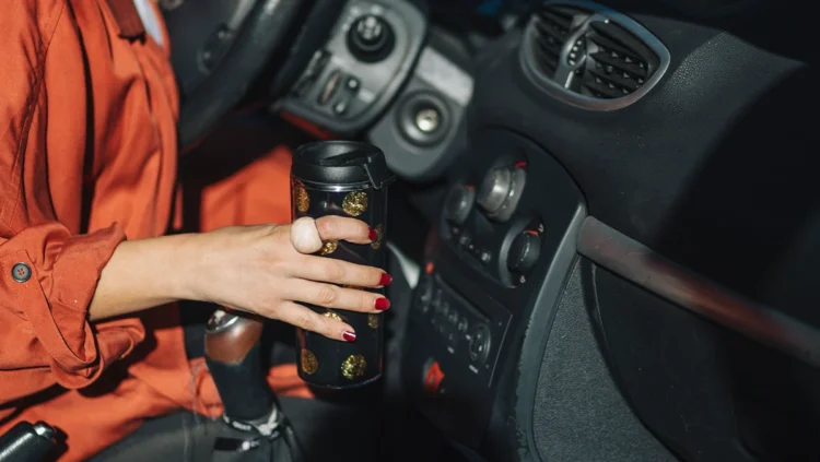 Motorista segurando um copo térmico no interior do carro, representando conforto, segurança e acessórios automotivos que facilitam o dia a dia ao dirigir.