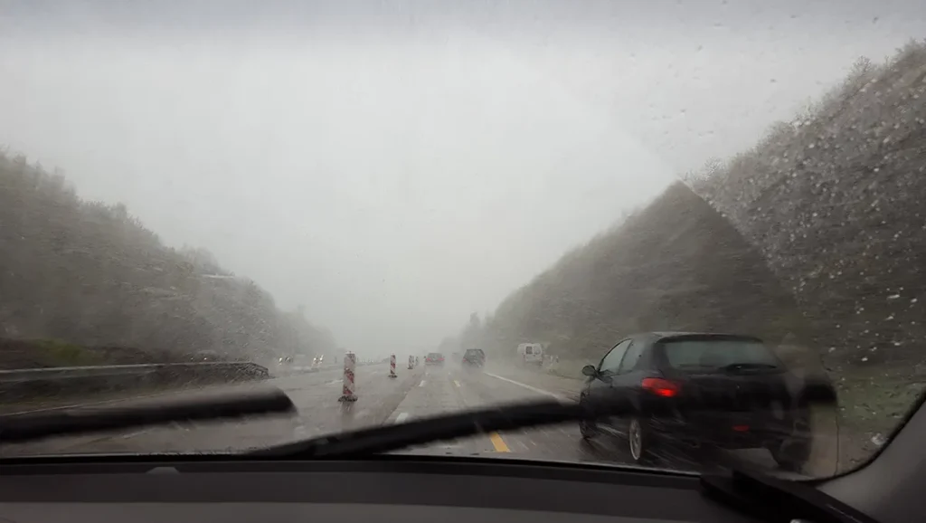 Vista de estrada sob forte tempestade, com chuva intensa e baixa visibilidade através do para-brisa do carro, simbolizando o alerta laranja do Inmet com ventos acima de 90 km/h e riscos no trânsito.