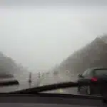 Vista de estrada sob forte tempestade, com chuva intensa e baixa visibilidade através do para-brisa do carro, simbolizando o alerta laranja do Inmet com ventos acima de 90 km/h e riscos no trânsito.