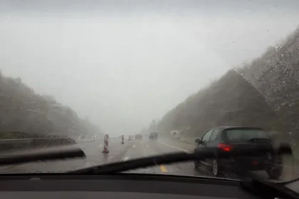 Vista de estrada sob forte tempestade, com chuva intensa e baixa visibilidade atrav&eacute;s do para-brisa do carro, simbolizando o alerta laranja do Inmet com ventos acima de 90 km/h e riscos no tr&acirc;nsito.