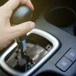 Mão mudando a marcha de um câmbio automático em um carro moderno, destacando o seletor de posições e botões de modo de condução, representando problemas e testes para identificar trancos no câmbio.