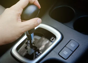 Mão mudando a marcha de um câmbio automático em um carro moderno, destacando o seletor de posições e botões de modo de condução, representando problemas e testes para identificar trancos no câmbio.