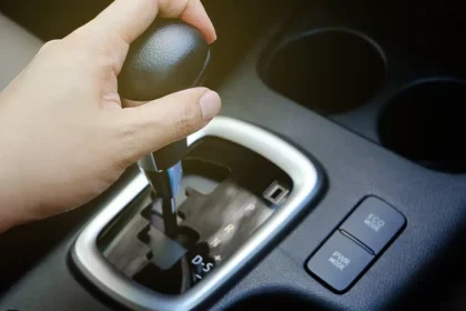 M&atilde;o mudando a marcha de um c&acirc;mbio autom&aacute;tico em um carro moderno, destacando o seletor de posi&ccedil;&otilde;es e bot&otilde;es de modo de condu&ccedil;&atilde;o, representando problemas e testes para identificar trancos no c&acirc;mbio.