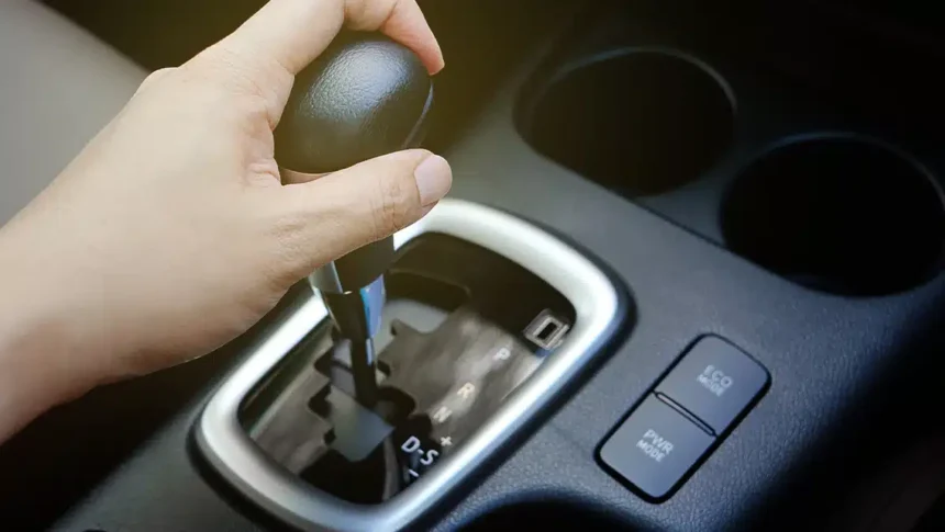 Mão mudando a marcha de um câmbio automático em um carro moderno, destacando o seletor de posições e botões de modo de condução, representando problemas e testes para identificar trancos no câmbio.