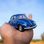 Mão segurando um Fusca azul em miniatura sob um céu azul, representando a tendência de transformar carros reais em versões digitais com aparência de miniatura para posts criativos e virais nas redes sociais.