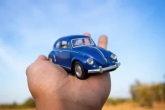 Mão segurando um Fusca azul em miniatura sob um céu azul, representando a tendência de transformar carros reais em versões digitais com aparência de miniatura para posts criativos e virais nas redes sociais.