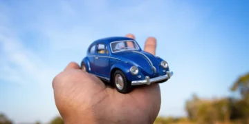 Mão segurando um Fusca azul em miniatura sob um céu azul, representando a tendência de transformar carros reais em versões digitais com aparência de miniatura para posts criativos e virais nas redes sociais.