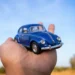 Mão segurando um Fusca azul em miniatura sob um céu azul, representando a tendência de transformar carros reais em versões digitais com aparência de miniatura para posts criativos e virais nas redes sociais.