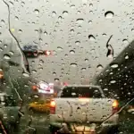 Para-brisa de carro coberto por gotas de chuva em estrada movimentada, com baixa visibilidade e luzes de veículos refletindo na pista molhada, ilustrando o alerta laranja do Inmet e os riscos de aquaplanagem.