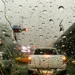 Para-brisa de carro coberto por gotas de chuva em estrada movimentada, com baixa visibilidade e luzes de veículos refletindo na pista molhada, ilustrando o alerta laranja do Inmet e os riscos de aquaplanagem.