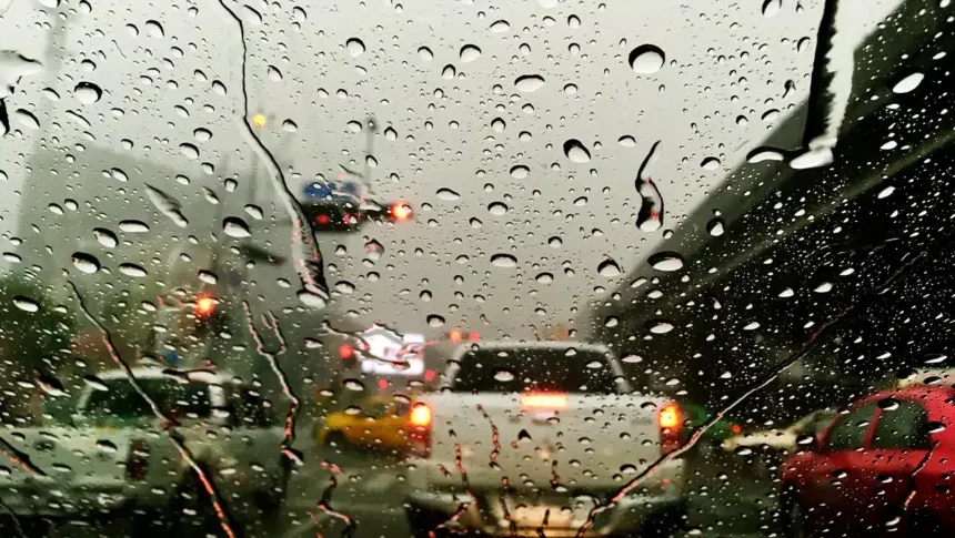 Chuva forte nas estradas: guia completo para dirigir com segurança no alerta laranja do Inmet e evitar aquaplanagem 1 Para-brisa de carro coberto por gotas de chuva em estrada movimentada, com baixa visibilidade e luzes de veículos refletindo na pista molhada, ilustrando o alerta laranja do Inmet e os riscos de aquaplanagem.