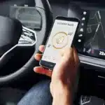 Motorista segura um smartphone com aplicativo de economia de energia enquanto o painel do carro exibe informações de navegação, ilustrando a integração entre conectividade automotiva, infotainment e telemetria nos veículos de 2025.