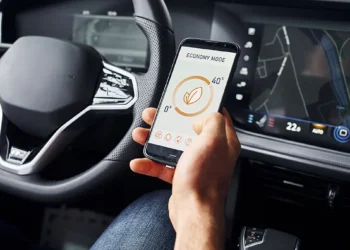 Motorista segura um smartphone com aplicativo de economia de energia enquanto o painel do carro exibe informações de navegação, ilustrando a integração entre conectividade automotiva, infotainment e telemetria nos veículos de 2025.