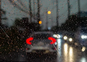 Vista de dentro de um carro em dia chuvoso, com gotas de água no para-brisa e veículos à frente com faróis acesos, representando os desafios e a importância da direção segura sob chuva.
