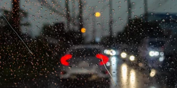 Vista de dentro de um carro em dia chuvoso, com gotas de água no para-brisa e veículos à frente com faróis acesos, representando os desafios e a importância da direção segura sob chuva.
