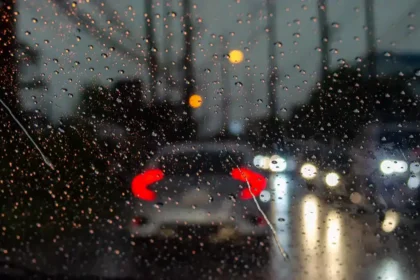 Vista de dentro de um carro em dia chuvoso, com gotas de &aacute;gua no para-brisa e ve&iacute;culos &agrave; frente com far&oacute;is acesos, representando os desafios e a import&acirc;ncia da dire&ccedil;&atilde;o segura sob chuva.