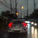 Vista de dentro de um carro em dia chuvoso, com gotas de água no para-brisa e veículos à frente com faróis acesos, representando os desafios e a importância da direção segura sob chuva.