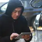 Criminoso de capuz usando um tablet para acessar o sistema eletrônico de um carro, representando o golpe do CAN Invader usado para invadir veículos sem chave.