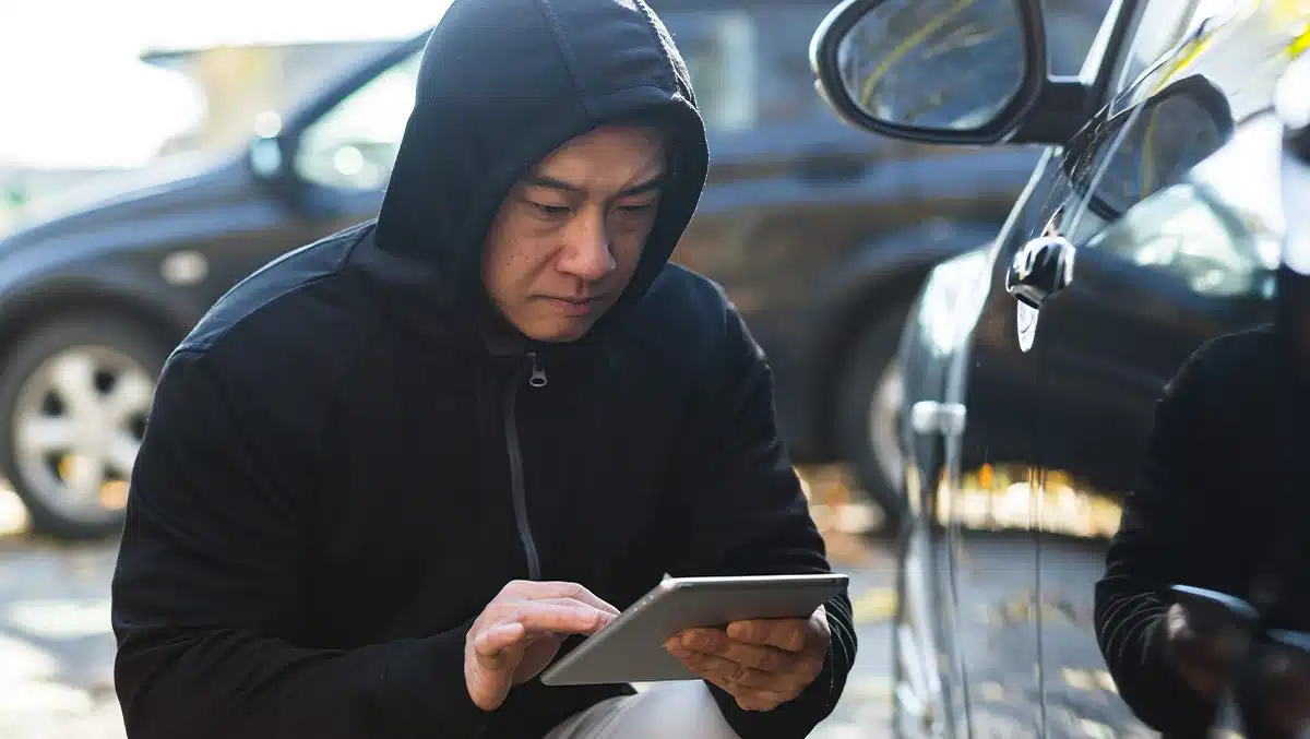 Criminoso de capuz usando um tablet para acessar o sistema eletrônico de um carro, representando o golpe do CAN Invader usado para invadir veículos sem chave.