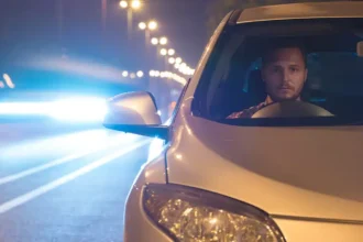 Motorista dirige à noite em rodovia iluminada, com o farol do carro ligado e outro veículo vindo em sentido oposto, representando a aplicação da Lei do Farol Automotivo e a importância da iluminação correta para segurança nas estradas.
