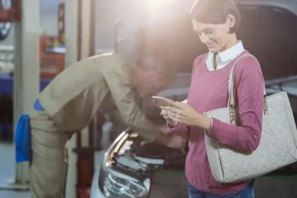 Mec&acirc;nico atendendo cliente em oficina moderna, com mulher segurando celular ao lado do carro, representando a nova era da manuten&ccedil;&atilde;o preventiva automotiva com foco em ve&iacute;culos el&eacute;tricos, h&iacute;bridos e conectados.