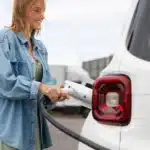 Mulher conectando cabo de recarga em carro elétrico branco em estação pública, representando o crescimento das vendas de veículos elétricos no Brasil e a expansão da infraestrutura de recarga.