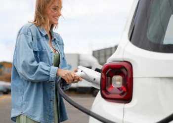 Mulher conectando cabo de recarga em carro elétrico branco em estação pública, representando o crescimento das vendas de veículos elétricos no Brasil e a expansão da infraestrutura de recarga.
