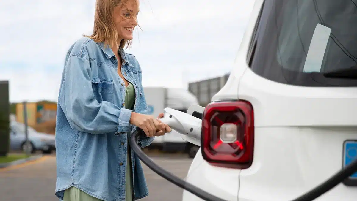 Mulher conectando cabo de recarga em carro elétrico branco em estação pública, representando o crescimento das vendas de veículos elétricos no Brasil e a expansão da infraestrutura de recarga.