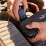 Mãos conectando carregador de bateria automotivo em motor de carro, mostrando o uso correto do equipamento para recarregar a bateria com segurança.