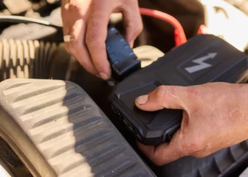 Mãos conectando carregador de bateria automotivo em motor de carro, mostrando o uso correto do equipamento para recarregar a bateria com segurança.