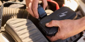 Mãos conectando carregador de bateria automotivo em motor de carro, mostrando o uso correto do equipamento para recarregar a bateria com segurança.