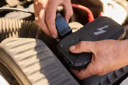 M&atilde;os conectando carregador de bateria automotivo em motor de carro, mostrando o uso correto do equipamento para recarregar a bateria com seguran&ccedil;a.