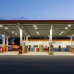 Imagem horizontal mostrando posto de combustíveis e oficina com sinais de atenção, fiscais em ação e elementos que representam os devedores contumazes câmara fecha