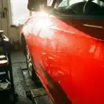 Carro com foco na porta, destacando riscos leves e microrelevo sob o sol, ferramentas de polimento ao lado na oficina