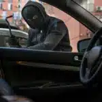 Homem mascarado tentando arrombar a porta de um carro estacionado em área urbana, representando o aumento dos furtos de veículos no Brasil em 2025.