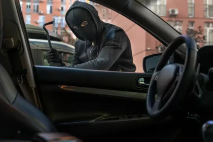 Homem mascarado tentando arrombar a porta de um carro estacionado em &aacute;rea urbana, representando o aumento dos furtos de ve&iacute;culos no Brasil em 2025.