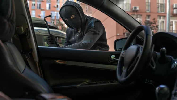 Homem mascarado tentando arrombar a porta de um carro estacionado em área urbana, representando o aumento dos furtos de veículos no Brasil em 2025.