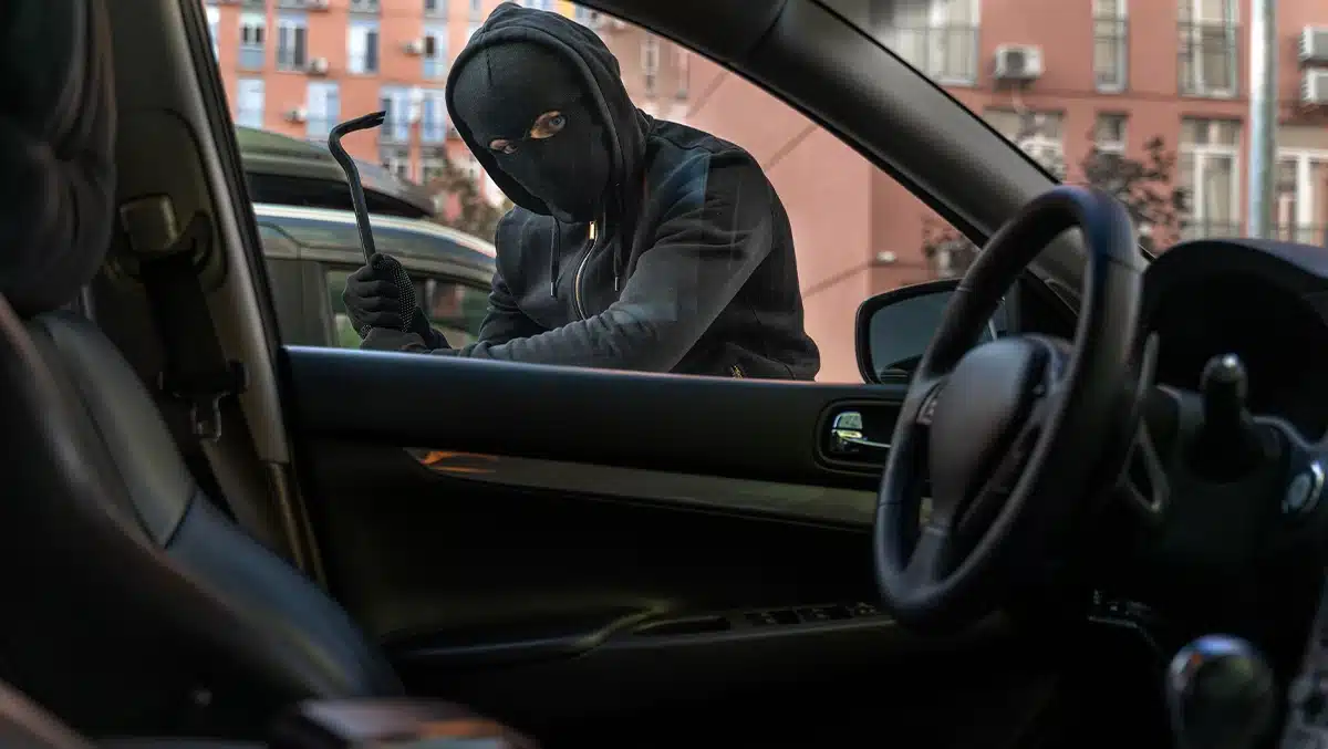 Homem mascarado tentando arrombar a porta de um carro estacionado em área urbana, representando o aumento dos furtos de veículos no Brasil em 2025.