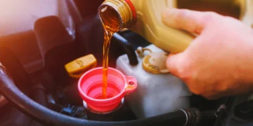 Mão de motorista despejando óleo de motor através de funil rosa em compartimento do carro durante a troca de óleo automotivo, representando manutenção preventiva e cuidados com o veículo.