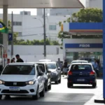 Posto de gasolina em Pernambuco com painel de preços alto, carros abastecendo, bandeiras brasileiras e movimento urbano