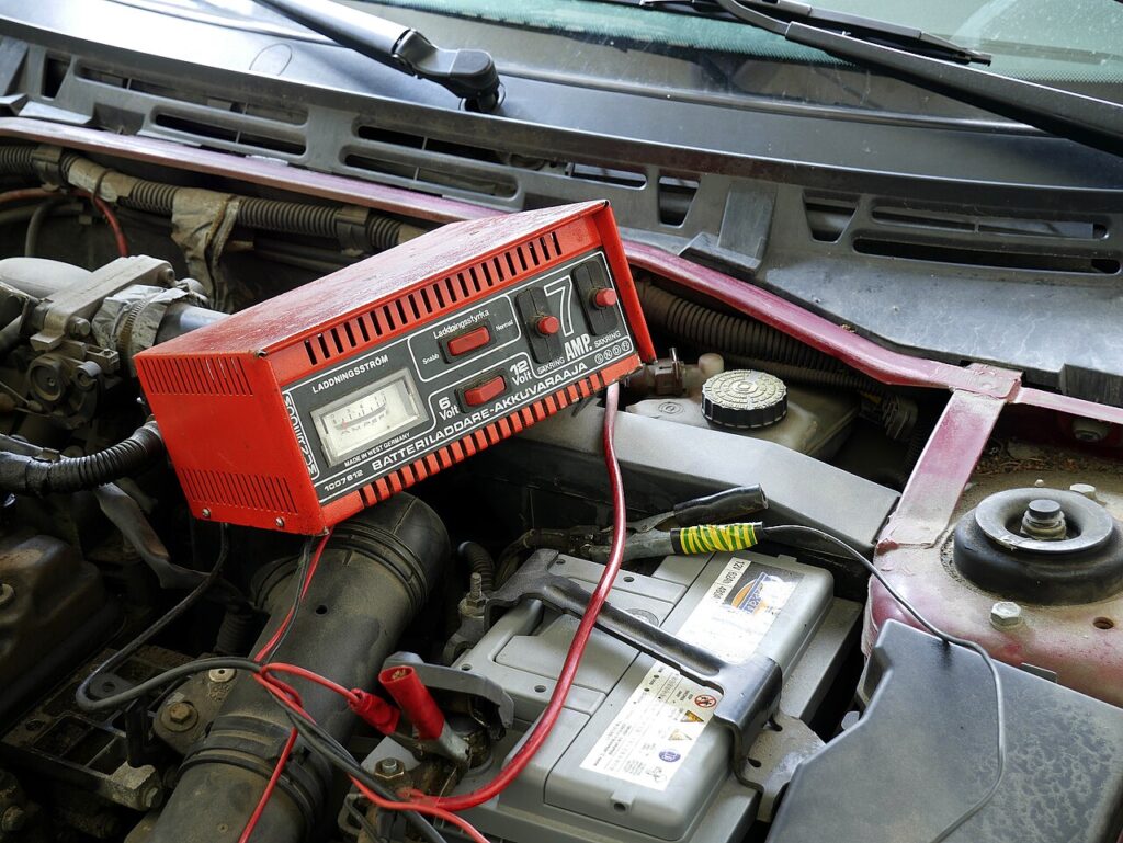 Como carregar bateria de carro no próprio veículo: carregador preso aos polos, capô aberto e ambiente ventilado.