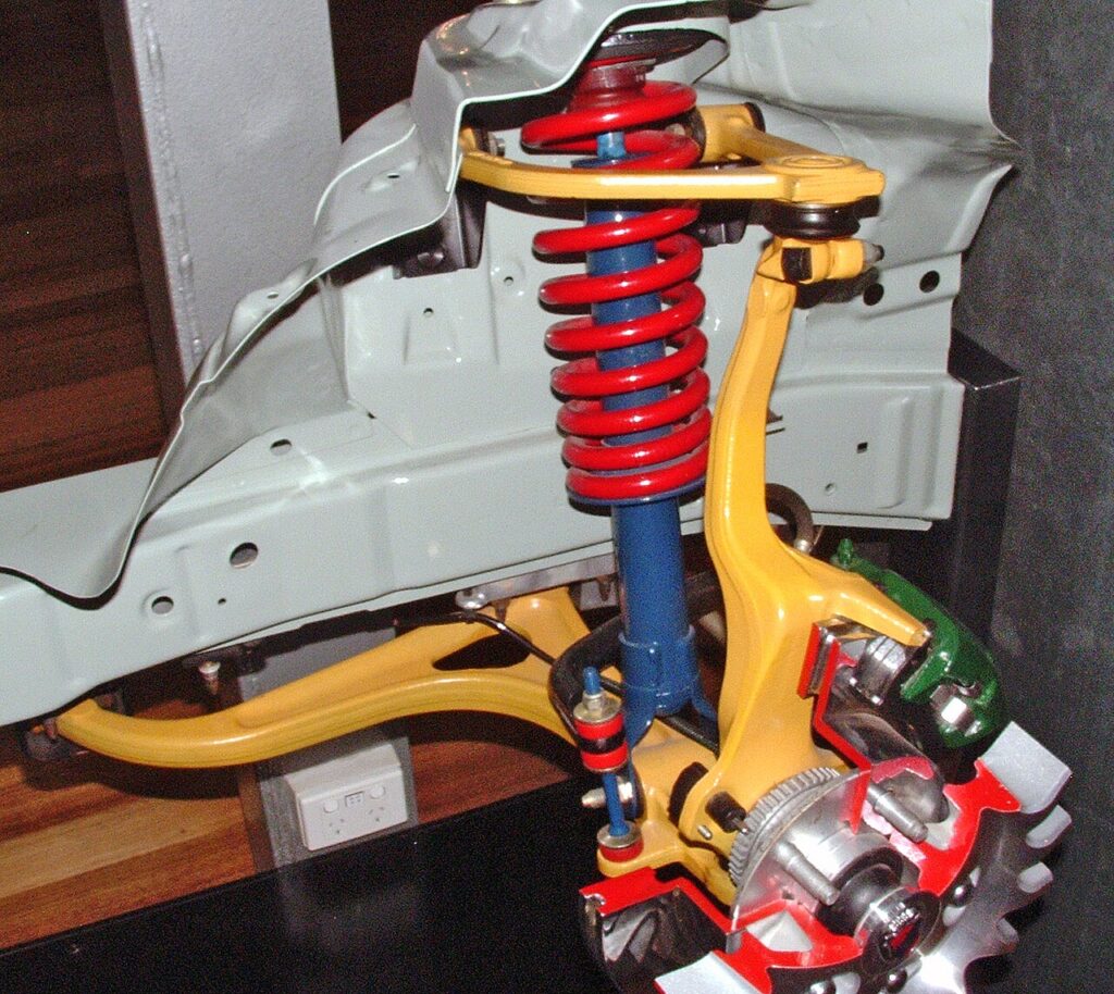 Suspensão de duplo triângulo (double wishbone): os braços pintados de amarelo ligam a roda à carroceria; mola e amortecedor ficam no meio.
