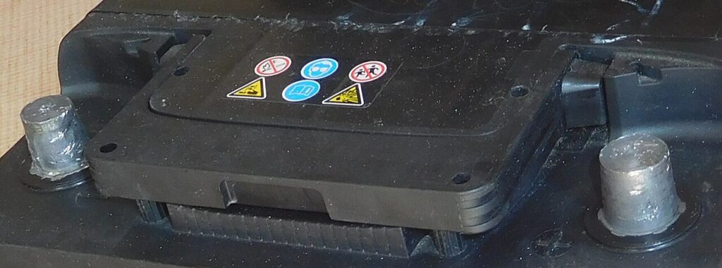 Troca de bateria de carro: passo a passo 2 Troca de bateria de carro: os dois polos ficam no alto da caixa; o negativo (−) é o primeiro a ser solto. Os símbolos lembram óculos, nada de chama e ácido.