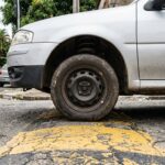 Barulho metálico na suspensão: roda dianteira de veículo passando por lombada em rua urbana de asfalto, mostrando a compressão da suspensão ao transpor a ondulação.