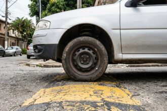 Barulho metálico na suspensão: roda dianteira de veículo passando por lombada em rua urbana de asfalto, mostrando a compressão da suspensão ao transpor a ondulação.