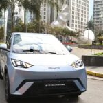 BYD Dolphin Mini: hatch compacto elétrico moderno conectado a ponto de recarga na rua de uma grande cidade brasileira durante o entardecer.