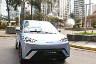 BYD Dolphin Mini: hatch compacto elétrico moderno conectado a ponto de recarga na rua de uma grande cidade brasileira durante o entardecer.