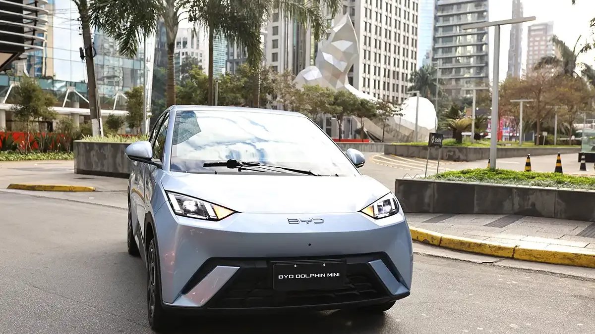 BYD Dolphin Mini: hatch compacto elétrico moderno conectado a ponto de recarga na rua de uma grande cidade brasileira durante o entardecer.