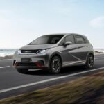 BYD Dolphin Mini: carro compacto elétrico conectado a cabo de carregamento em rua arborizada com prédios urbanos ao fundo.