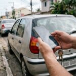 Consulta de placa de veículo: mãos de motorista segurando celular perto da traseira de um carro estacionado em rua brasileira, simulando consulta de dados.